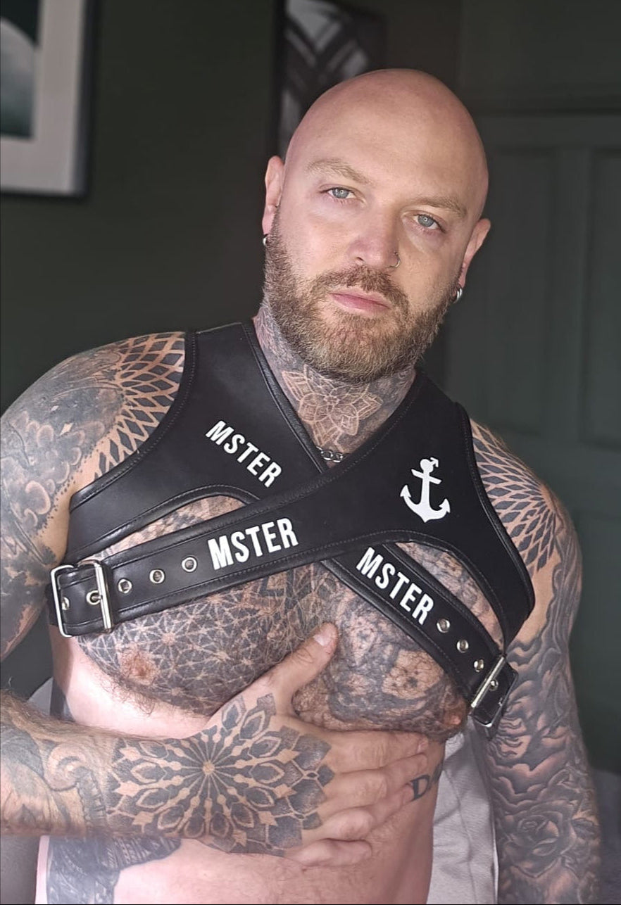 Man wearing a leather harness with 'MSTER' branding, surrounded by tattoos.