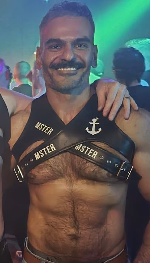 Man wearing a leather harness with 'MSTER' branding at a party.