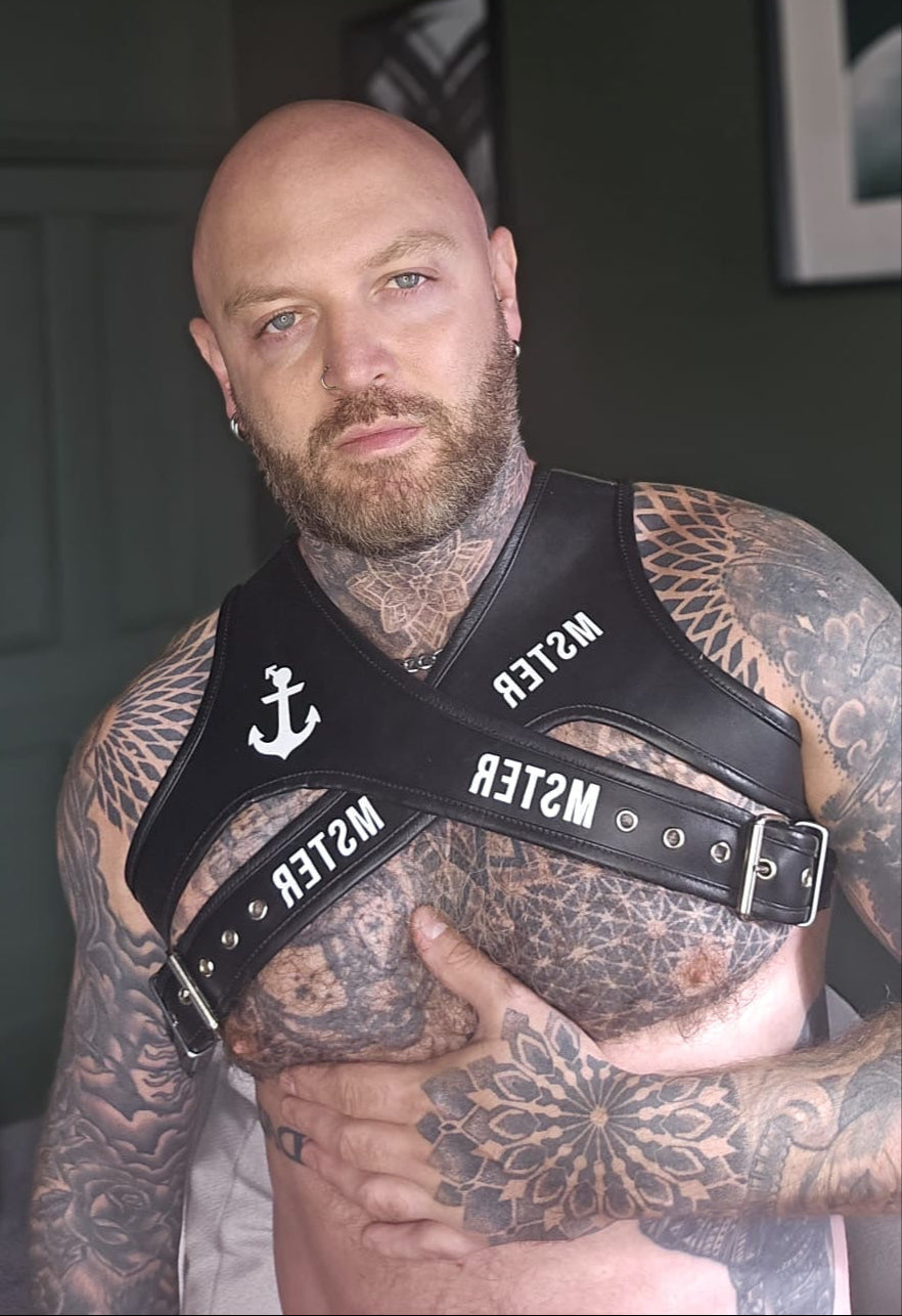 Man wearing a leather harness with 'MSTER' branding, surrounded by tattoos.