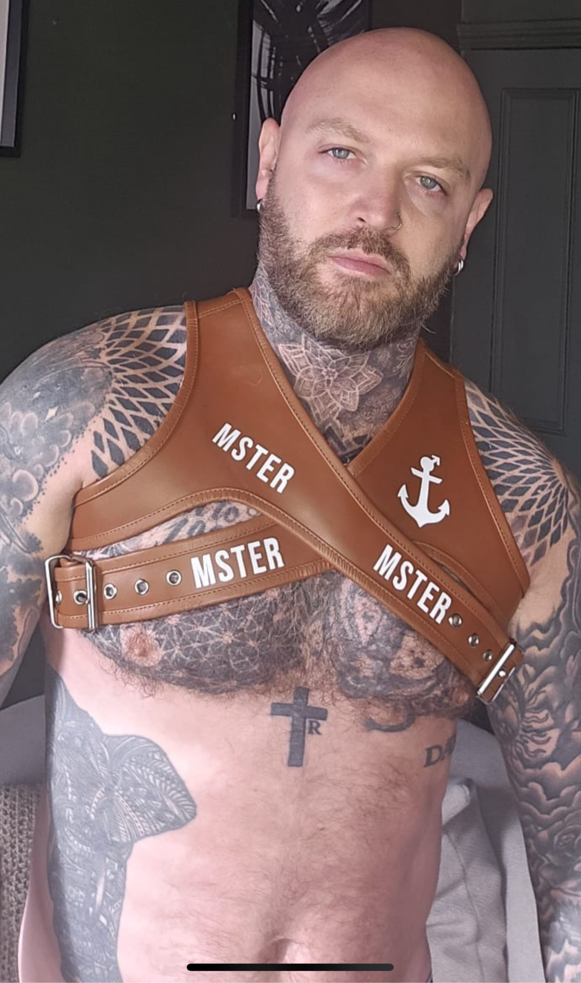 Man wearing a leather harness with 'MSTER' branding, tattoos visible on arms and chest.